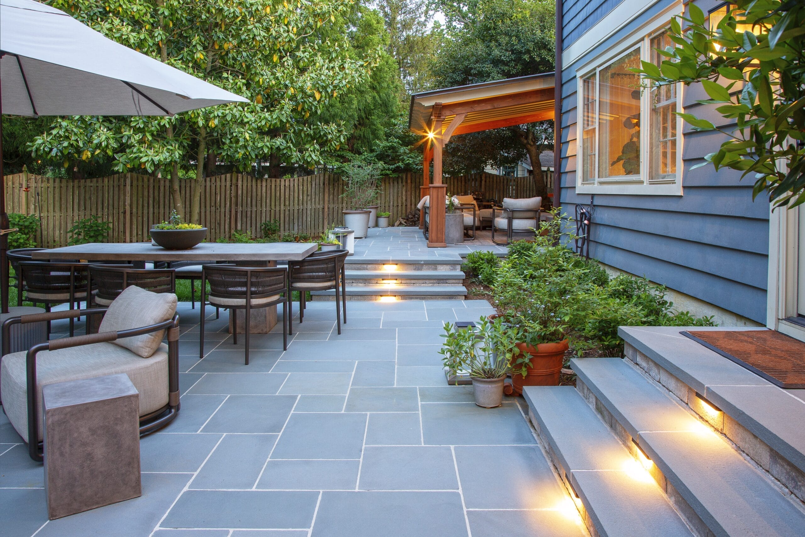 Blue-gray stone patio with illuminated steps, outdoor dining area, pergola, and modern seating - Ridge
