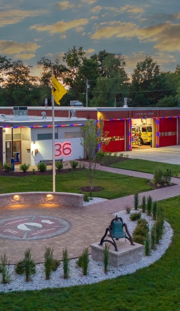 Fire station courtyard with Ridge circular paver patio, memorial design, landscaped beds, and fire bell monument at dusk