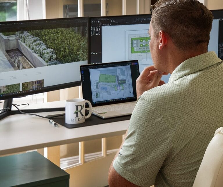 Landscape designer at Ridge Studios reviewing digital garden plans on laptop and dual monitors, with Ridge Studios mug on desk.