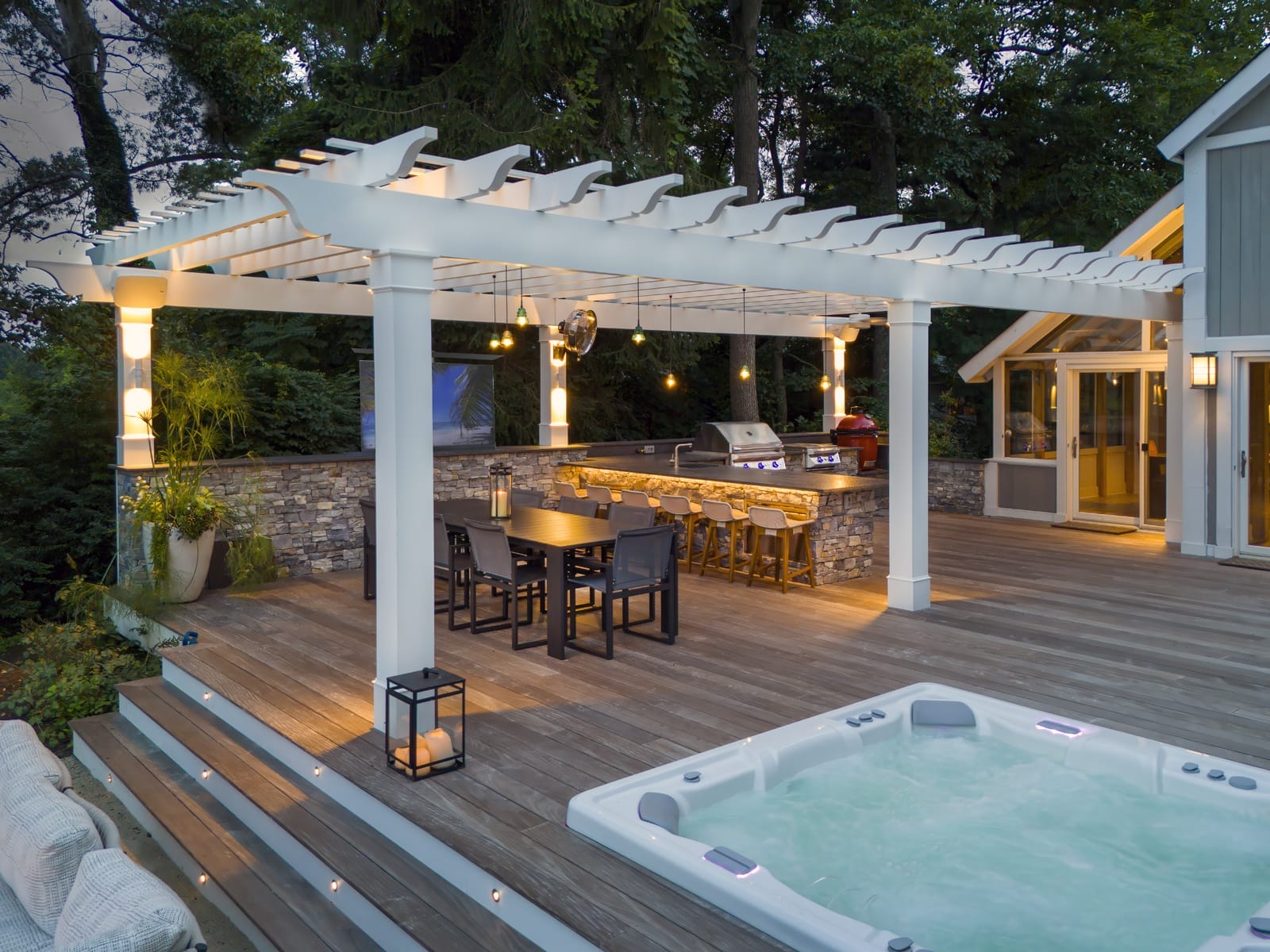 Ridge outdoor living space with white pergola, string lights, stone bar with seating, outdoor kitchen, composite deck, and hot tub.